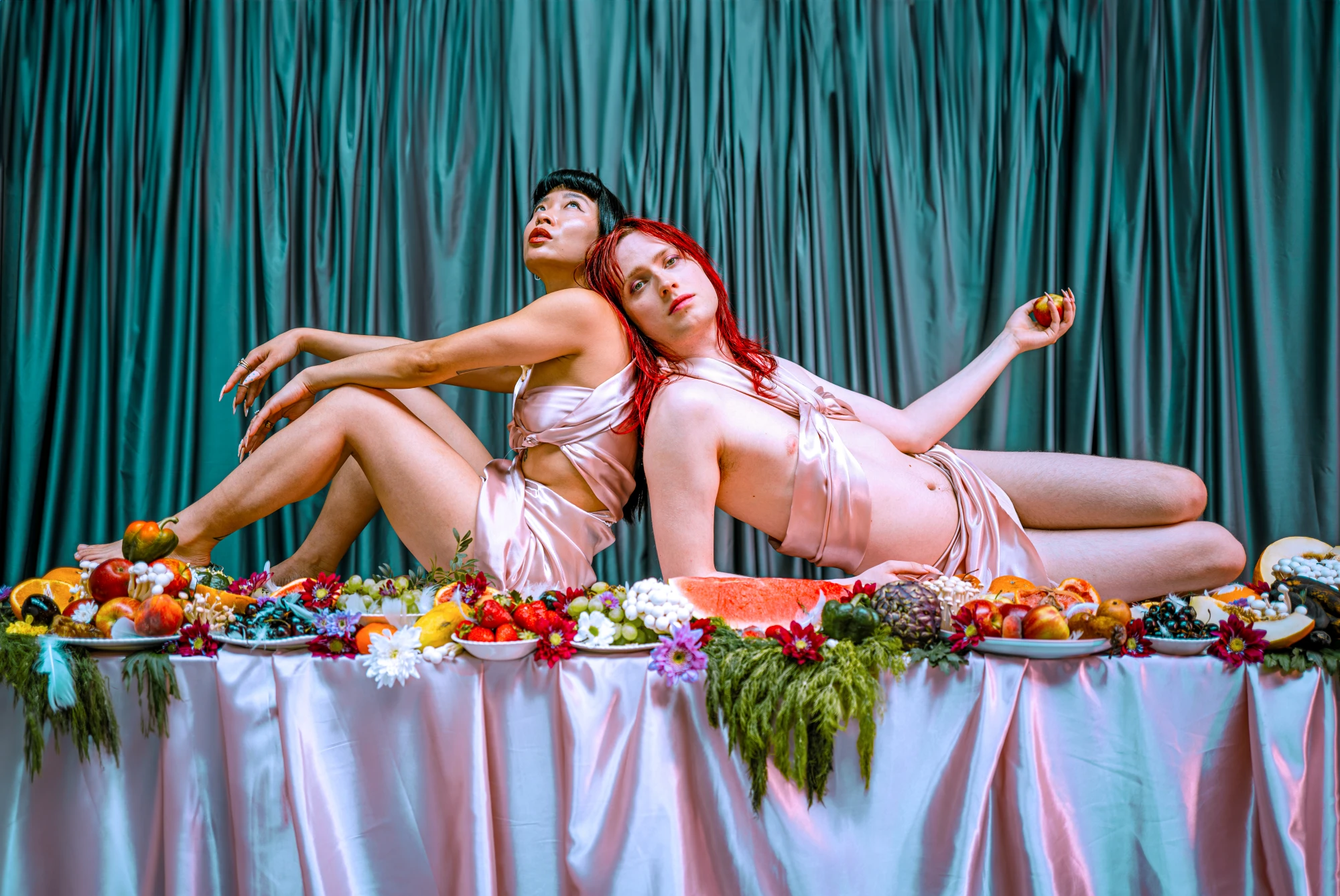 A woman and a non binary femme figure hold fruit whilst on top of a table covered in fruit.