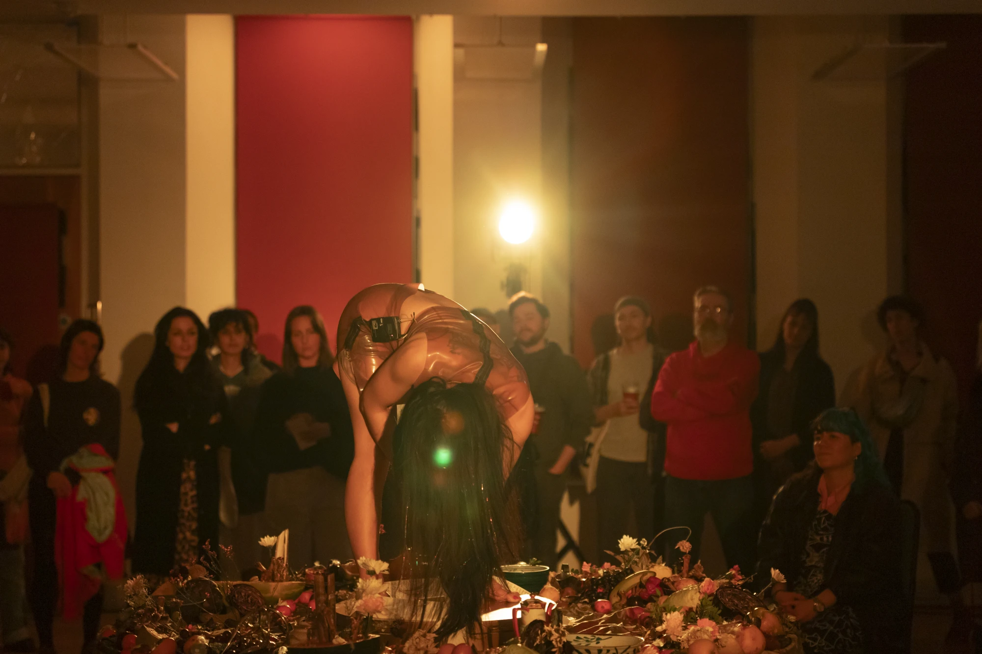 performance documentation of a woman with long black hair bent over on top of a circular table covered in rotting fruit, whilst wearing see through latex. The crowd watches her.
