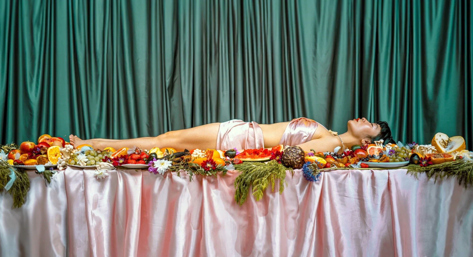 a woman lies across a table covered in rotting fruit.