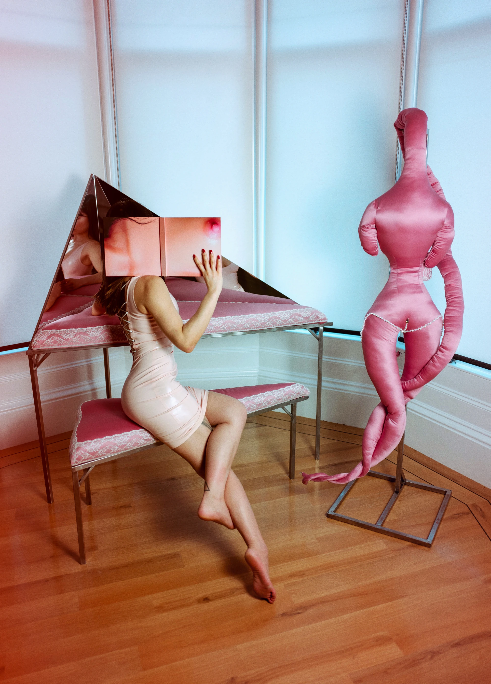 a woman holds a book over her face, as she sits in front of a triangle dressng table next to a soft satin sculpture that mimicks a feminine form.