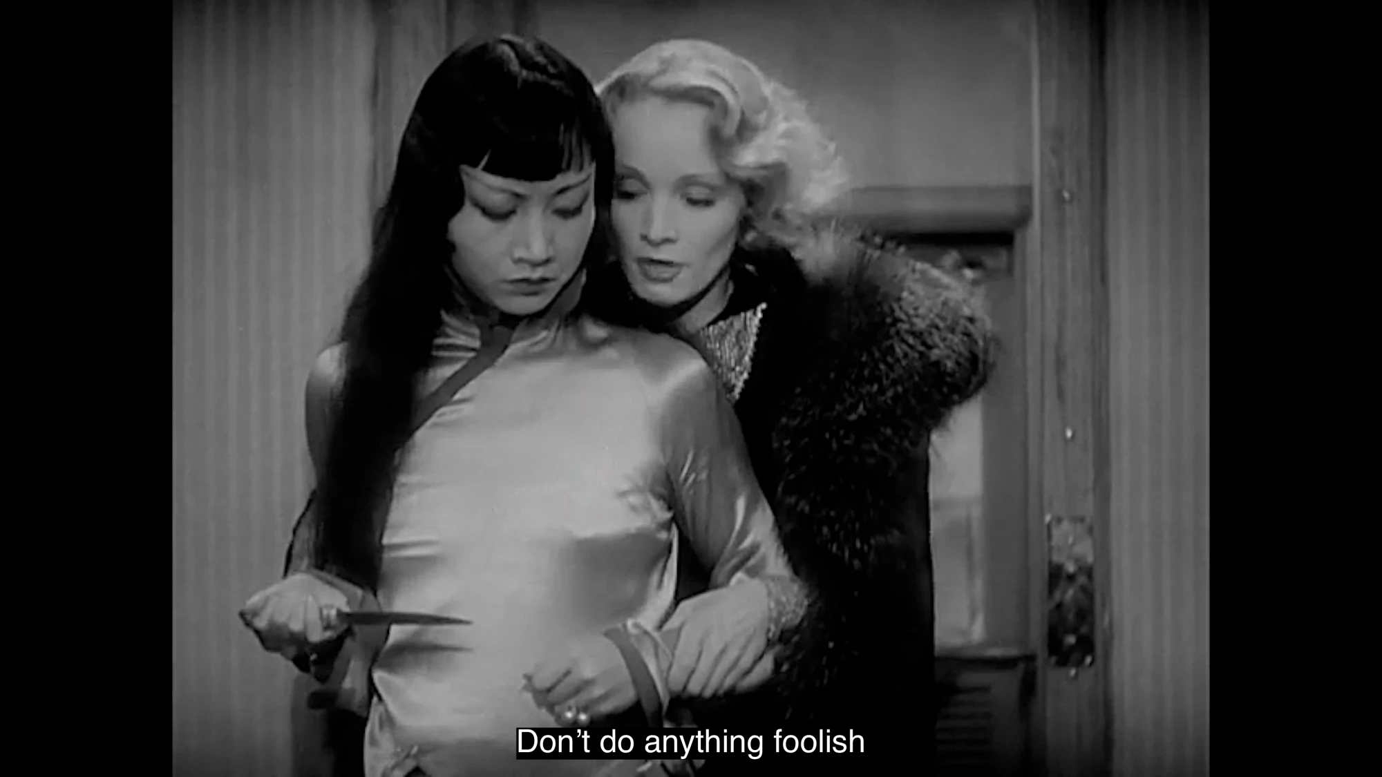A black and white film still featuring an Asian woman holding a knife, and a white woman behind her speaking into her ear. The caption reads 'Don't do anything foolish'.