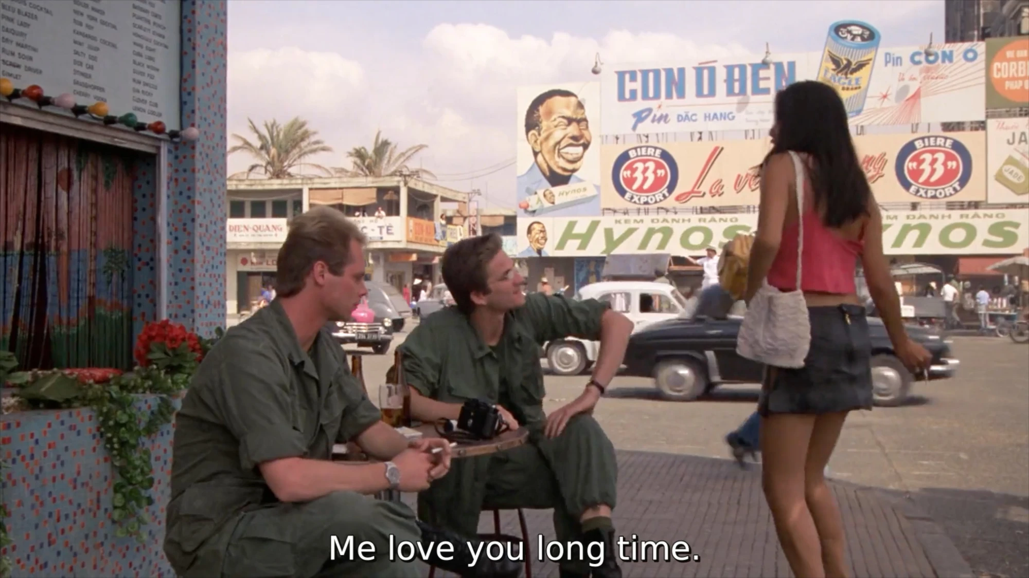 A film still featuring an Asian woman with long black hair and a miniskirt approaching two soldiers sitting on the side of the street. The caption reads 'Me love you long time'.