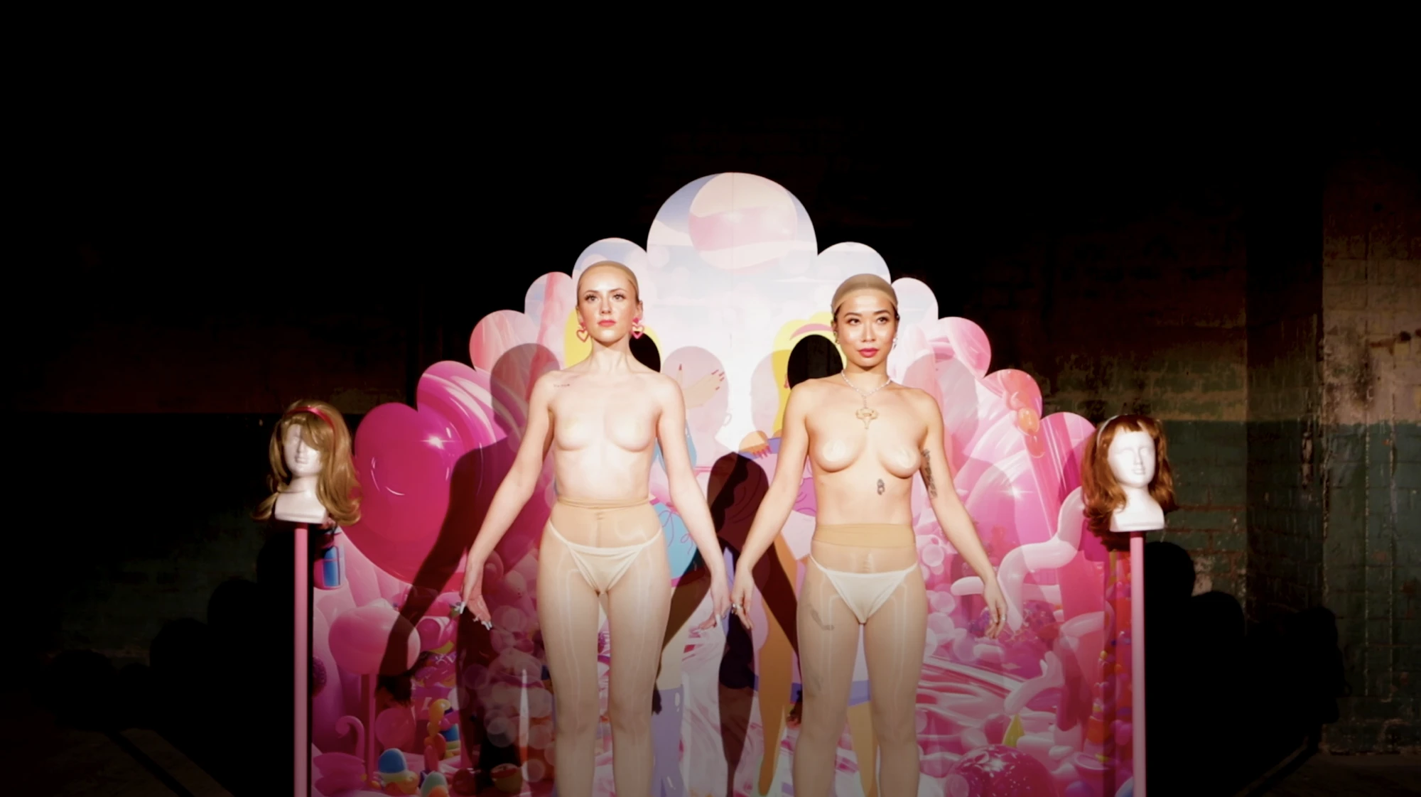 two woman srtand in front of a set, wearing flesh coloured tights and a flesh coloured skull cap.