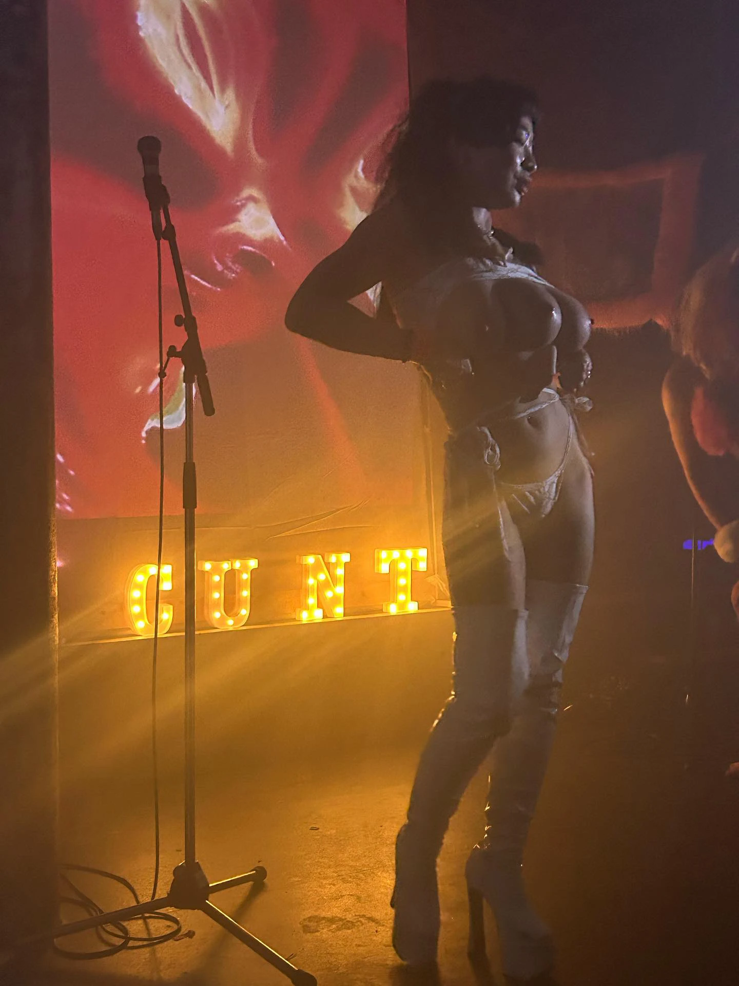 a woman with three breasts stand in front of light up lights that read 'cunt'.