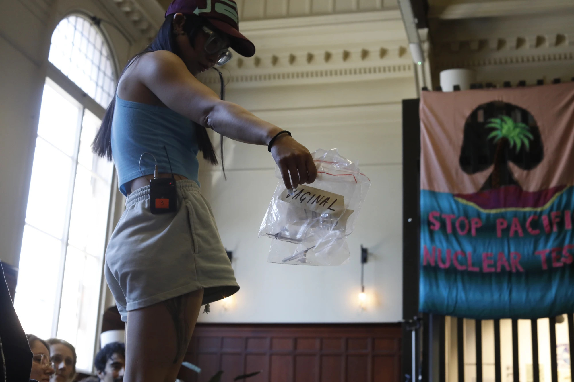 A chinese woman in her mid thirties is wearing a blue t shirt and a cap. She wears thick glasses and holds out a plastic bag that says 'VAGINAL'.