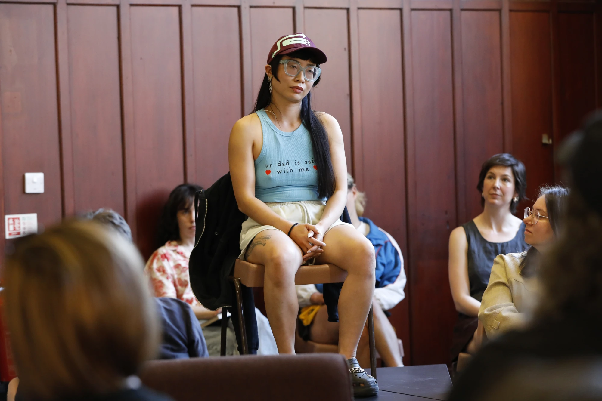 A chinese woman in her mid thirties is wearing a t shirt that says 'Your dad is safe with me', and a cap. She wears thick glasses and sits on an elevated chair in front of an audience.