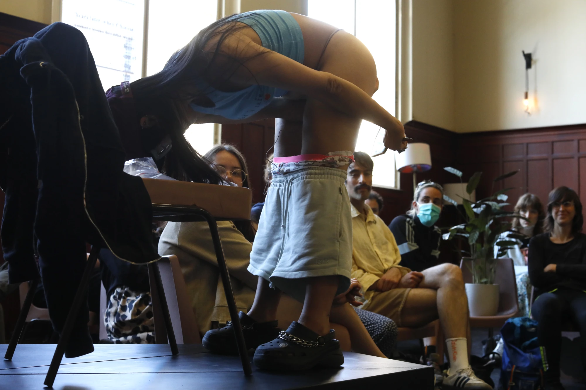 A chinese woman in her mid thirties is wearing a blue t shirt and a cap. She wears thick glasses and bends over on an elevated platform as she swabs herself while the audience watches.