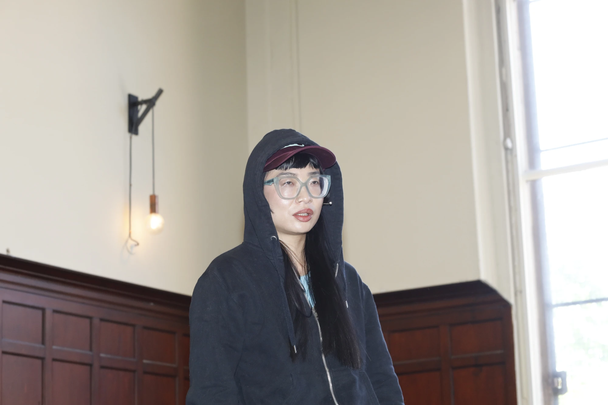 A chinese woman in her mid thirties is wearing a hoodie and a cap, with thick glasses.