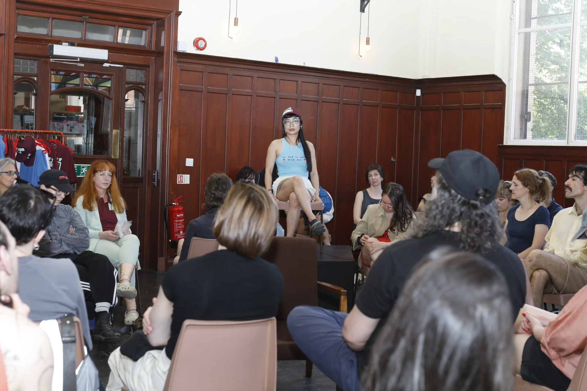 A chinese woman in her mid thirties is wearing a t shirt that says 'Your dad is safe with me', and a cap. She wears thick glasses and sits on an elevated chair in front of an audience.