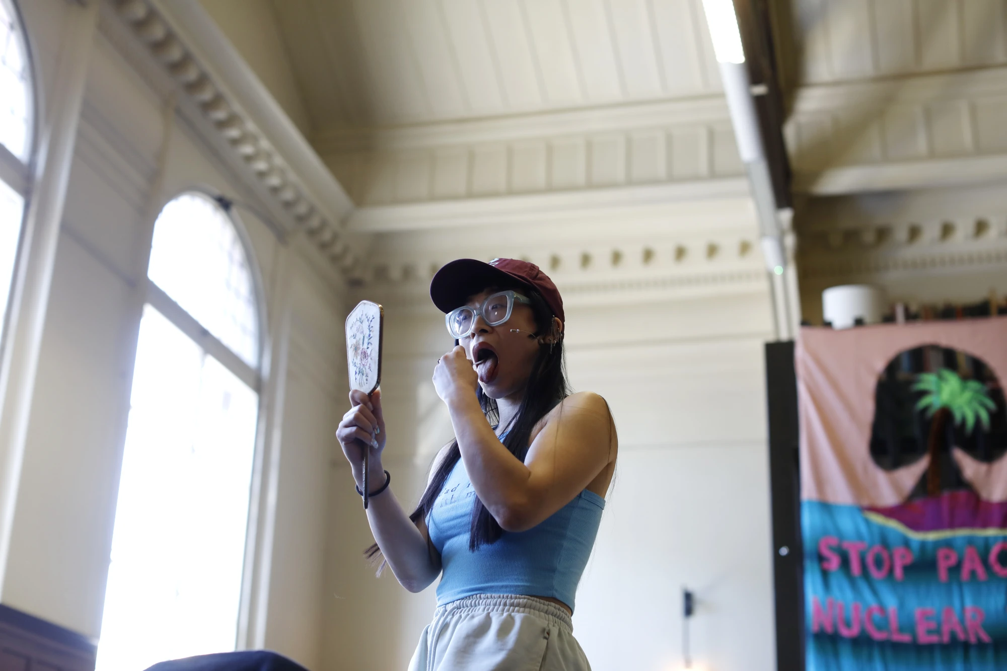 A chinese woman in her mid thirties is wearing a blue t shirt and a cap. She wears thick glasses and stands on an elevated platform swabbing her mouth with a hand held mirror.