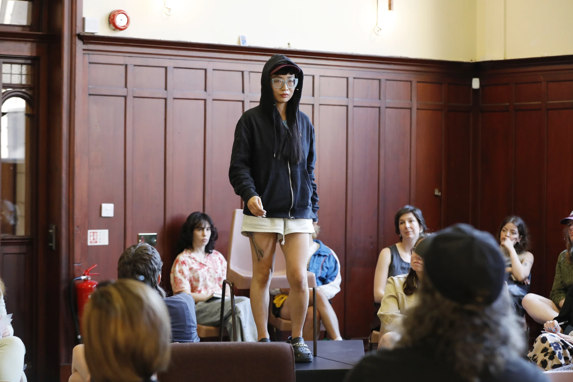 A chinese woman in her mid thirties is wearing a hoodie and a cap, with thick glasses. She walks on an elevated platform in front of seated audience.