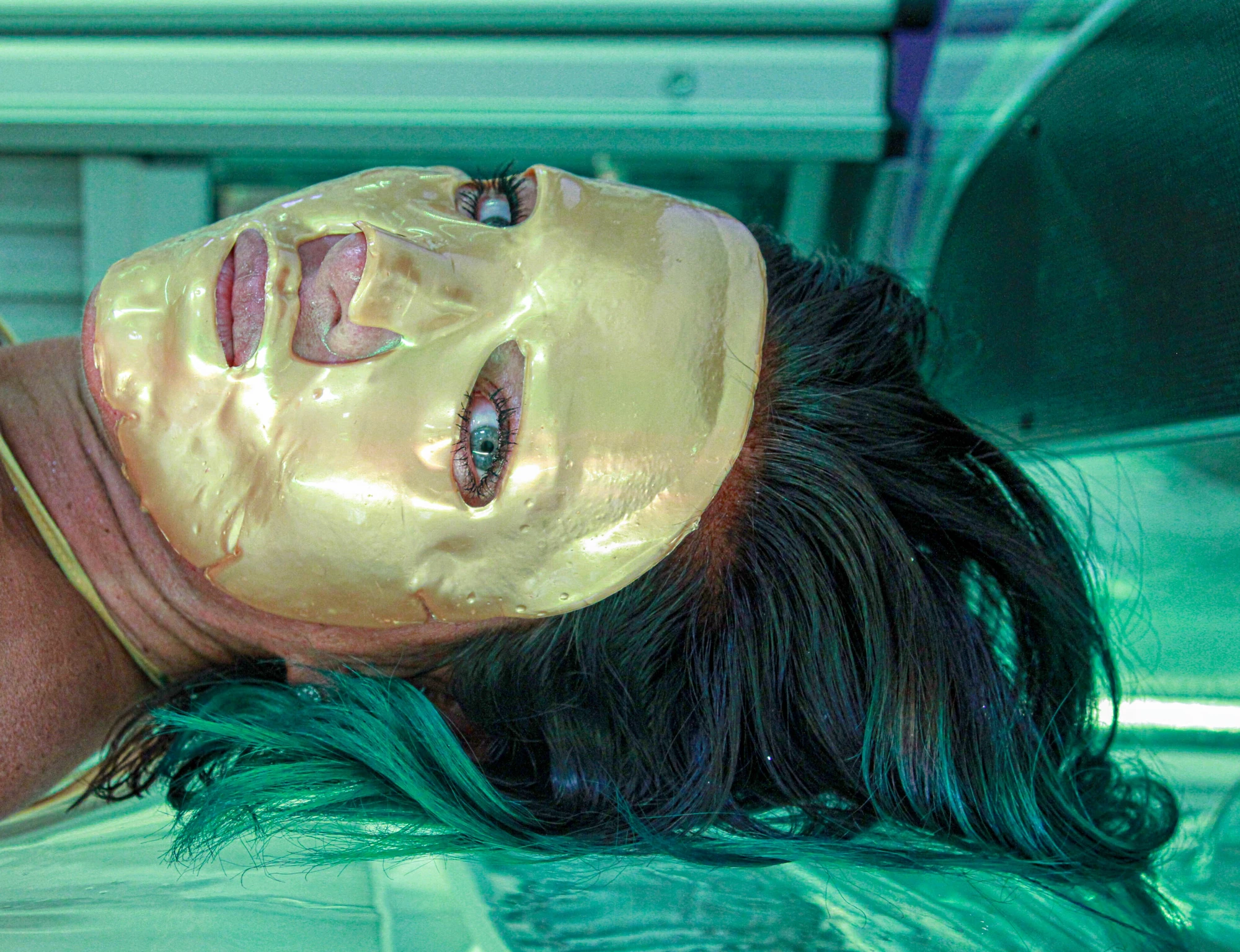 a woman is in a tanning booth with a gold face mask on, staring straight into the camera