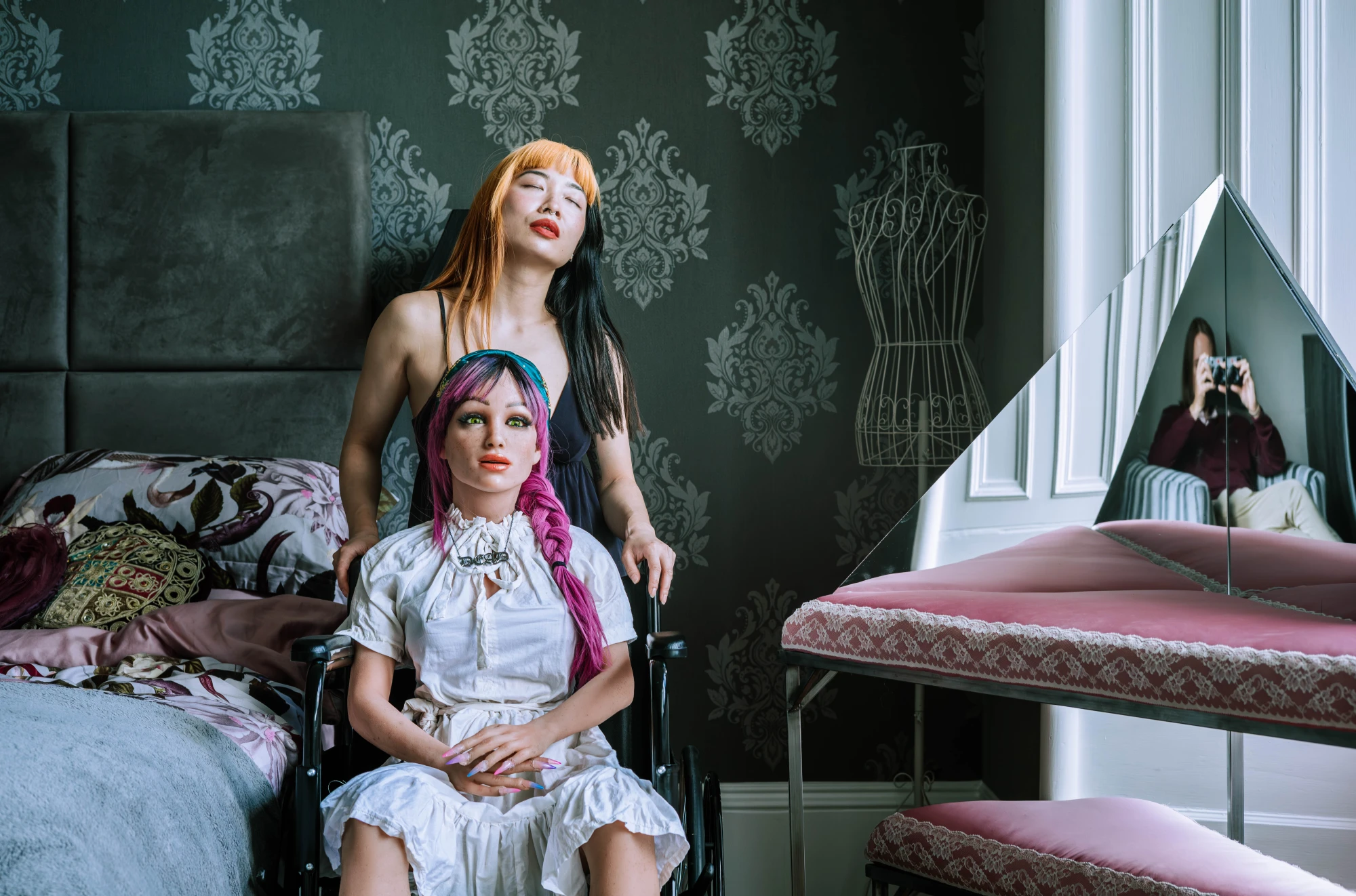A silicone love dolls sits on a wheelchair and a woman is standing behind her.