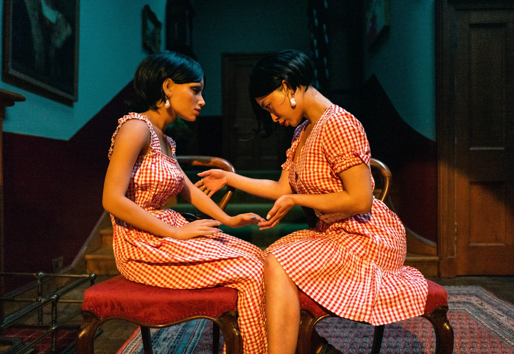 a silcone love doll sits in the hallway of a grand building facing a flesh and blood woman, who is examining the doll's hand. They are wearing matching red and white checkered dresses.