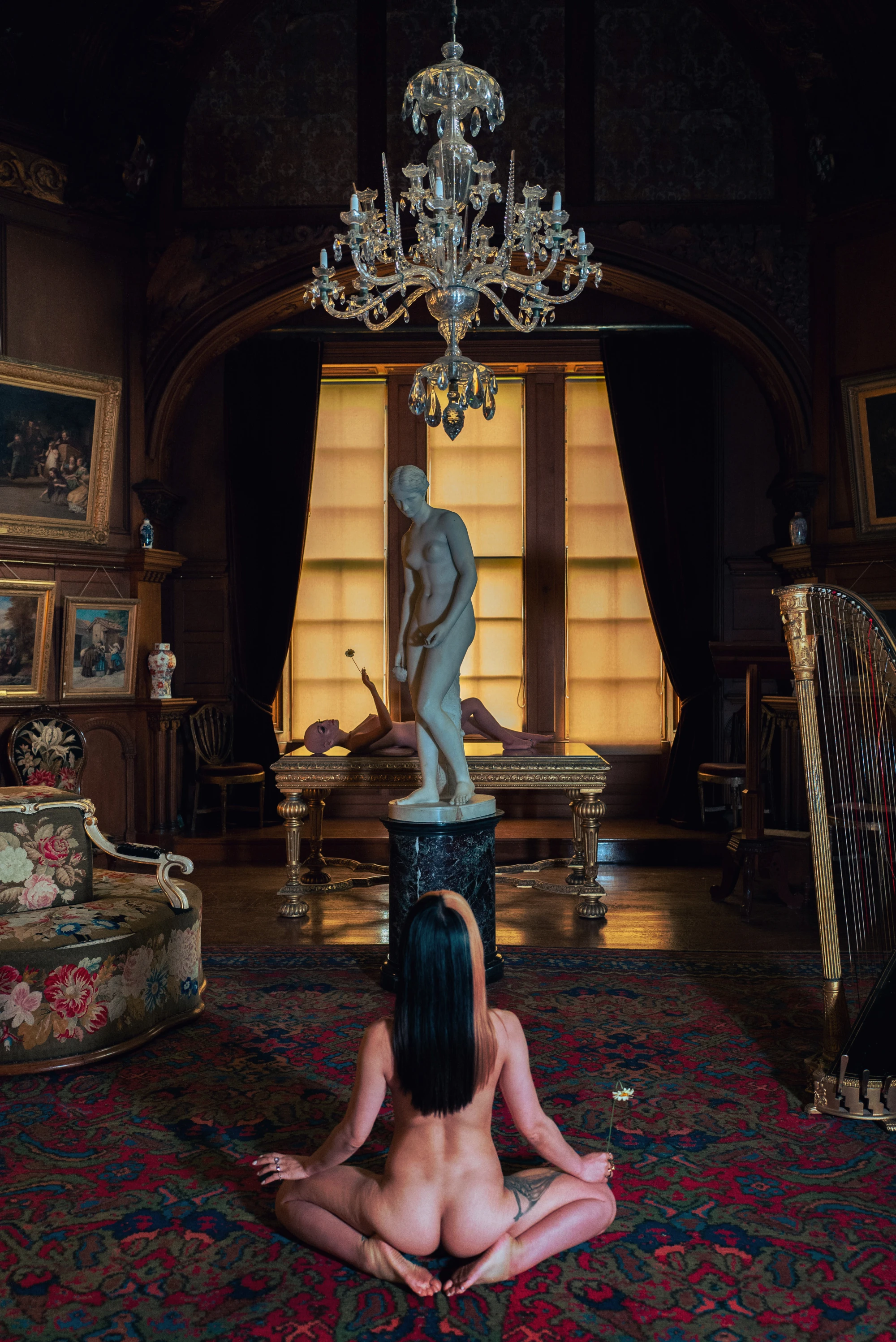 a woman sits in front of a statue. in the distance, a silicone love doll lies on a table.