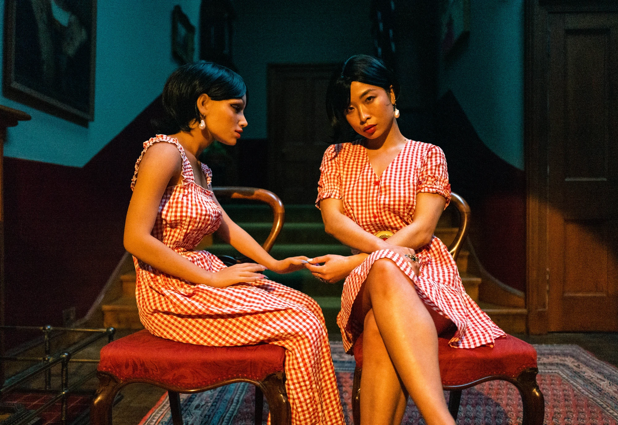 a silcone love doll sits in the hallway of a grand building facing a flesh and blood woman, who is holding her hand whilst looking inquisitively at the camera. They are wearing matching red and white checkered dresses.